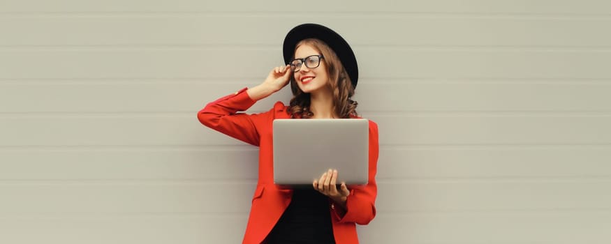 Happy modern young woman working on a laptop, freelancer girl with computer looking away in red suit, black hat against gray wall background