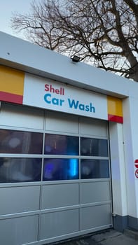 22.03.2025 Germany: Modern Shell Car Wash Facility With Blue Light Interior and Glass Doors. Shell car wash seen from the exterior, showcasing tinted glass doors and illuminating blue light visible inside, with surrounding architecture and trees emphasizing a modern and professional cleaning service.