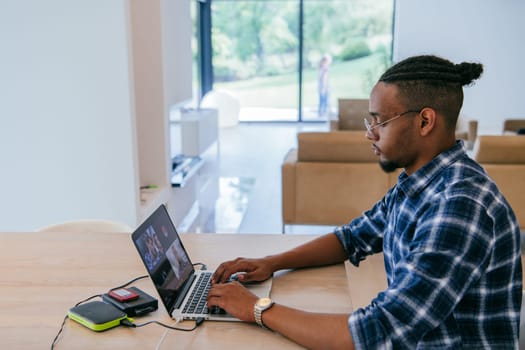 African American influencer in glasses sitting at a table in a modern living room, using a laptop for business video chat, conversation with friends and entertainment