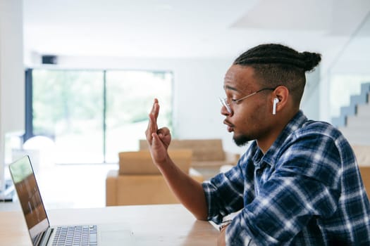 African American influencer in glasses sitting at a table in a modern living room, using a laptop for business video chat, conversation with friends and entertainment
