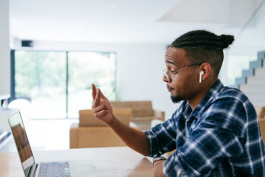 African American influencer in glasses sitting at a table in a modern living room, using a laptop for business video chat, conversation with friends and entertainment