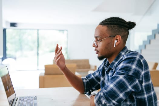 African American influencer in glasses sitting at a table in a modern living room, using a laptop for business video chat, conversation with friends and entertainment