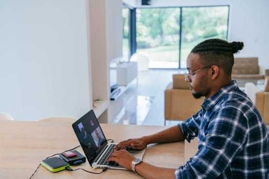 African American influencer in glasses sitting at a table in a modern living room, using a laptop for business video chat, conversation with friends and entertainment