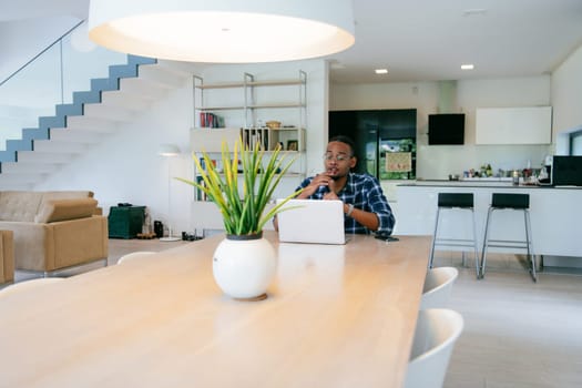 African American influencer in glasses sitting at a table in a modern living room, using a laptop for business video chat, conversation with friends and entertainment