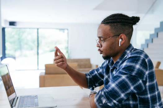 African American influencer in glasses sitting at a table in a modern living room, using a laptop for business video chat, conversation with friends and entertainment