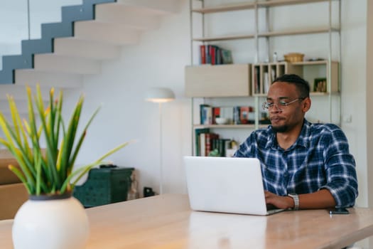 African American influencer in glasses sitting at a table in a modern living room, using a laptop for business video chat, conversation with friends and entertainment
