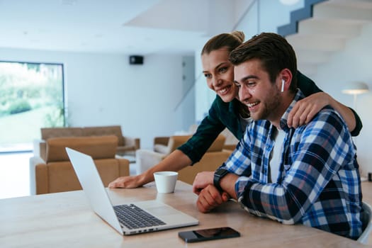 A young married couple is talking to parents, family and friends on a video call via a laptop while sitting in the living room of their modern house