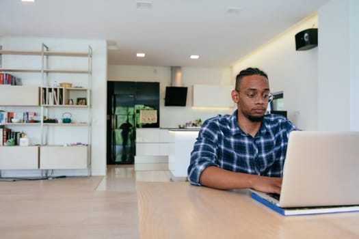 African American influencer in glasses sitting at a table in a modern living room, using a laptop for business video chat, conversation with friends and entertainment