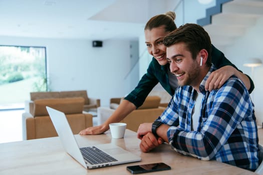 A young married couple is talking to parents, family and friends on a video call via a laptop while sitting in the living room of their modern house