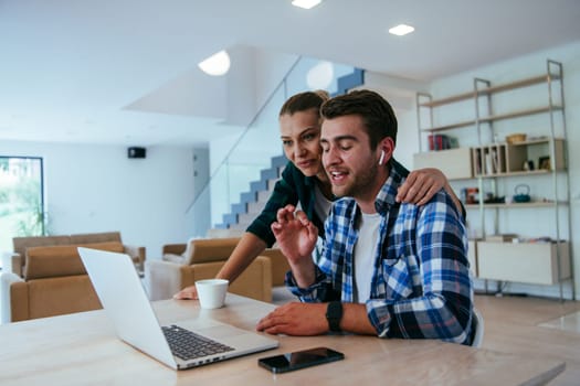 A young married couple is talking to parents, family and friends on a video call via a laptop while sitting in the living room of their modern house