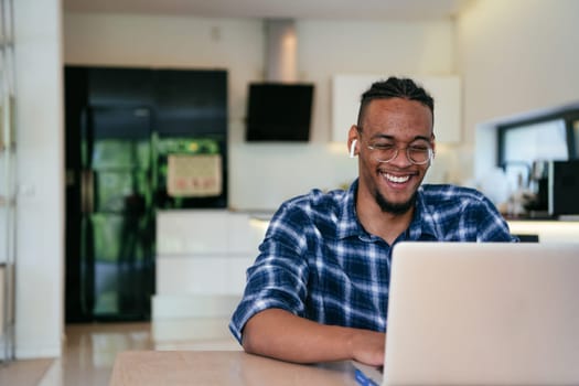 African American influencer in glasses sitting at a table in a modern living room, using a laptop for business video chat, conversation with friends and entertainment