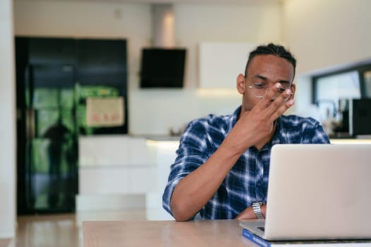 African American influencer in glasses sitting at a table in a modern living room, using a laptop for business video chat, conversation with friends and entertainment