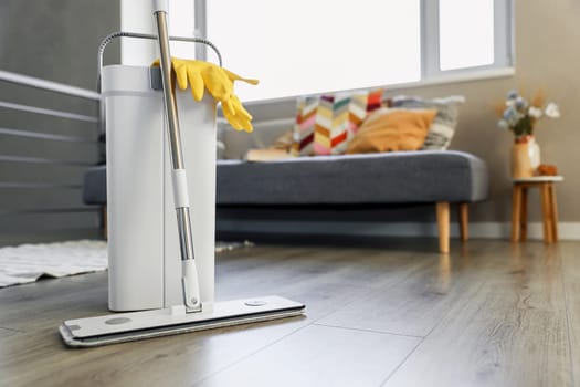 Cleaning supplies: mop, gloves, bucket with wringer sitting on wooden laminate floor in modern living room, wet equipment tools, housekeeping concept. Emphasizing home sanitation, cleanliness, hygiene