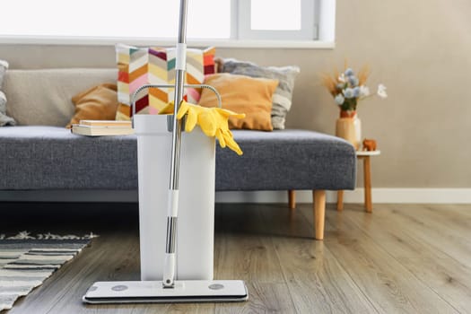 Cleaning supplies: mop, gloves, bucket with wringer sitting on wooden laminate floor in modern living room, wet equipment tools, housekeeping concept. Emphasizing home sanitation, cleanliness, hygiene