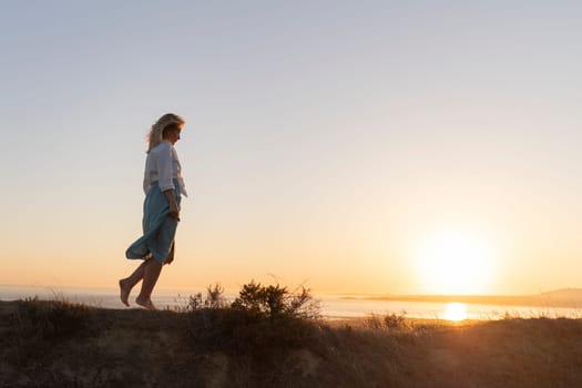 Mature woman walking barefoot on hilltop at sunset, enjoying freedom and connection with nature