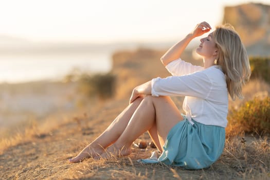 Blonde woman sits on a hilltop at sunset, embracing the tranquility of nature