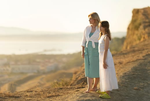 Mother and daughter standing barefoot on a hilltop, enjoying a breathtaking sunset over a coastal landscape