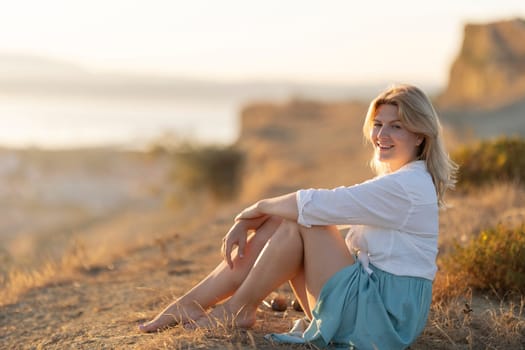 Blond woman is sitting on dry grass on a hilltop, enjoying the sunset over the sea