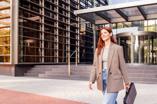 smiling young adult woman in casual suit walking in front of an office building holding a briefcase, concept of entrepreneur and business, copyspace for text