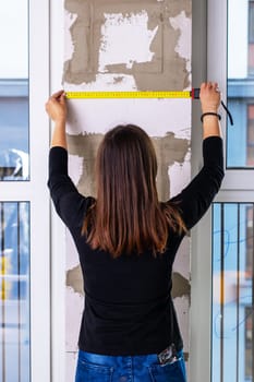 A woman diligently measures a walls dimensions using a tape measure, ensuring accuracy for her upcoming project