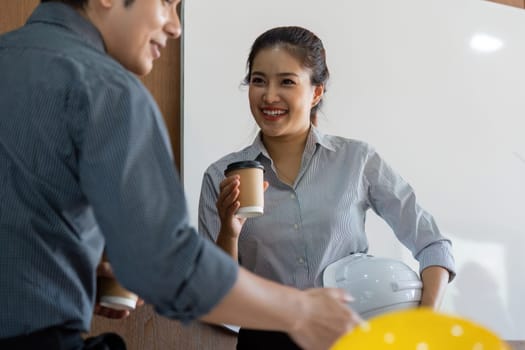 An engineer and colleague share a light moment over coffee, planning their next project amidst blueprints and a bright office setting. This scene captures teamwork and professionalism.