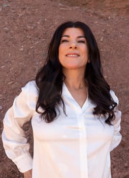 Portrait Of Smiling with Teeth Lebanese Woman In Her 50 in White Shirt Outside, Red Rocks On Background. Beautiful Confident Fashionable Female Outdoor, Middle Eastern Attractive Person. Vertical.