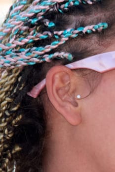 Close-up of a woman's ear adorned with a small, round earring. Her dreadlocks, in shades of blue, pink, and blonde, are styled and secured with a pink ribbon, highlighting their vibrant colors and textures.