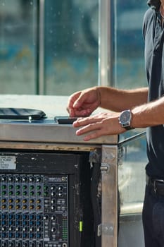 Catania. Italy - March 26, 2025: A person dressed in a black shirt operates a mixing console equipped with various knobs and inputs. Nearby, there is a smartphone and a small black case.