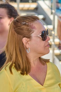 Portrait of a blonde woman with eyeglasses and colorful earrings, dressed in bright yellow. Outdoor setting with blurred natural elements.