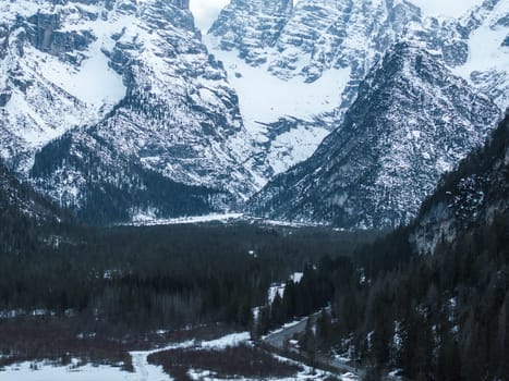 A frozen lake in the Alps is surrounded by dense evergreen forests and towering snow covered peaks. A winding road leads towards the serene landscape.