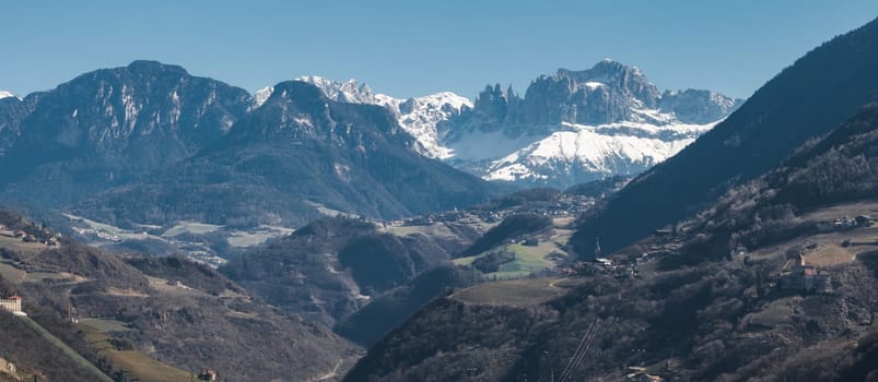 Aerial view of the Dolomites' snow capped peaks near Bolzano, Italy, with rolling hills, villages, vineyards, and a church in the foreground.