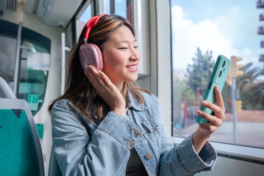 Young Chinese woman with pink headphones enjoying music on tram, eyes closed in relaxation. She holds her smartphone while leaning back in her seat, urban vibe and peaceful moment.