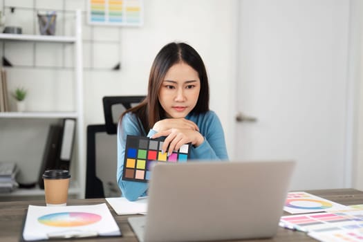 A designer thoughtfully reviews color samples while working on her laptop, indicating a focus on creating an impactful visual project.