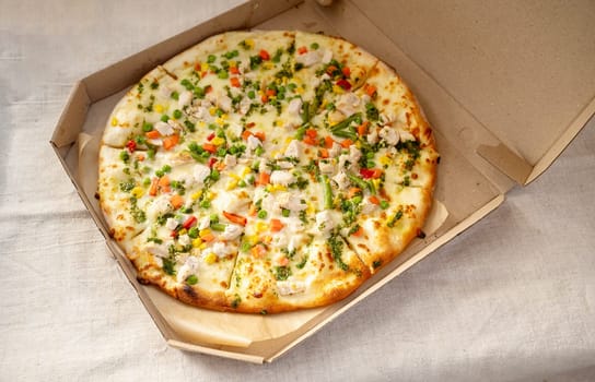 Delicious pizza with mozzarella, chicken, pepper, corn and green peas and carrots. vegetable pizza with chicken in a cardboard box of the delivery service. selective focus. linen tablecloth background