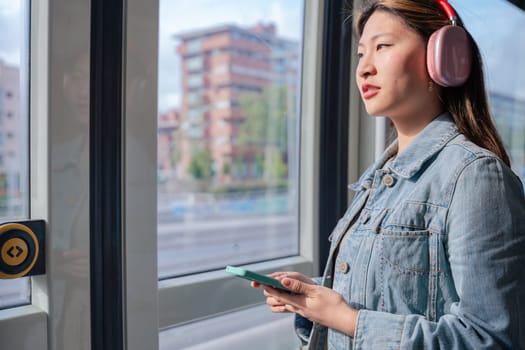 Young chinese woman wearing headphones and denim jacket holding smartphone while looking out the window of a city tram.Peaceful moment during commute, listening to music or thinking calmly.copy space