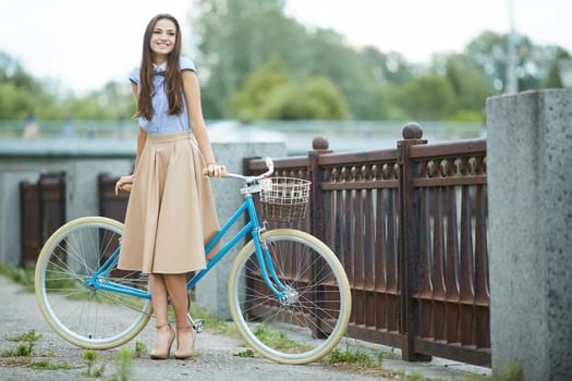 Stylish young woman with long hair smiling, posing with a vintage blue bicycle on a city bridge, wearing an elegant beige skirt and heels