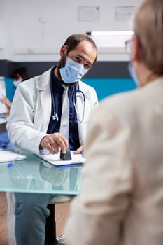 Young general practitioner in face mask is using stamper to complete prescription paper for female pensioner patient. Senior woman patiently waits as young doctor stamps medical paperwork in clinic.