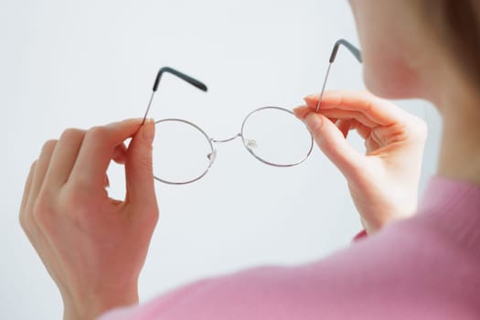 A smiling woman in a pink sweater holds a pair of round glasses towards the camera, set against a light blue background.