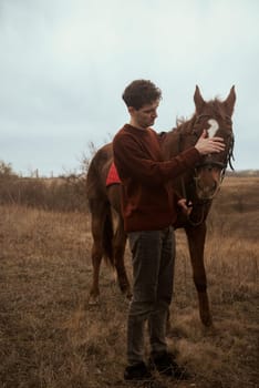 A man in a brown sweater is resting after horse riding with his horse. He is stroking his brown horse. Muchina and horse against the background of nature and a field with autumn grass. Concept of horse riding outdoors, sport and recreation.
