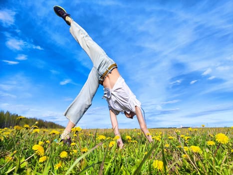 A joyful individual balances in a handstand on a colorful dandelion-filled meadow on a gorgeous and sunny day.