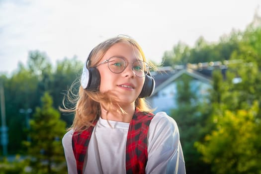 A young girl listens to music while standing outside, wearing headphones and smiling in a vibrant, sunny environment.