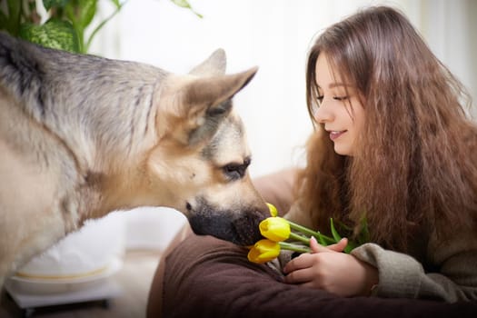 A woman with curly hair sits beside her dog, holding yellow tulips and sharing a moment in a warm, inviting home setting.