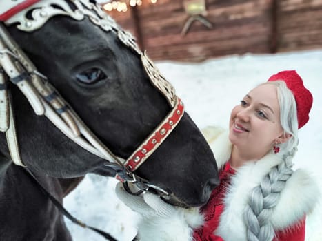 A joyful girl in traditional Russian attire shares a moment with a horse during the Maslenitsa holiday in winter.