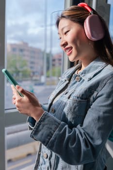 Young chinese woman wearing headphones and denim jacket holding smartphone while looking out the window of a city tram.Peaceful moment during commute, listening to music or thinking calmly.Vertical