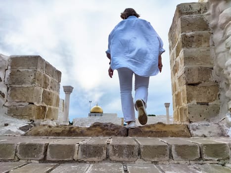 A middle-aged blonde woman walks up steps at a historic site, capturing the moment in a selfie while enjoying her travel excursion.