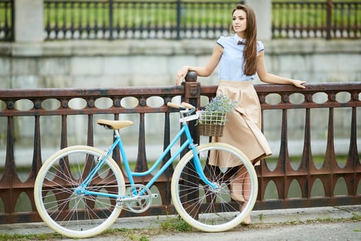 Stylish young woman with long hair smiling, posing with a vintage blue bicycle on a city bridge, wearing an elegant beige skirt and heels