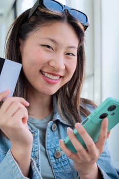 Happy chinese woman on tram holding a credit card and smartphone. Wearing sunglasses and denim jacket, smiling while shopping online or making a mobile payment during urban travel looking at camera.