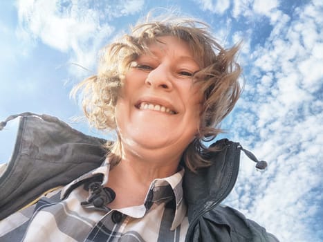 A middle-aged woman shares a joyful selfie under a bright blue sky with fluffy clouds while traveling.