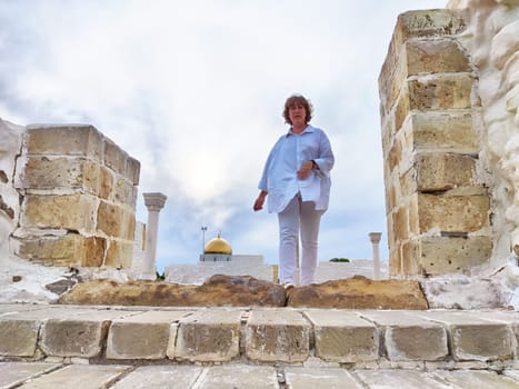 Blonde woman on a sightseeing excursion walks through ancient stone structures, enjoying a travel adventure under cloudy skies.