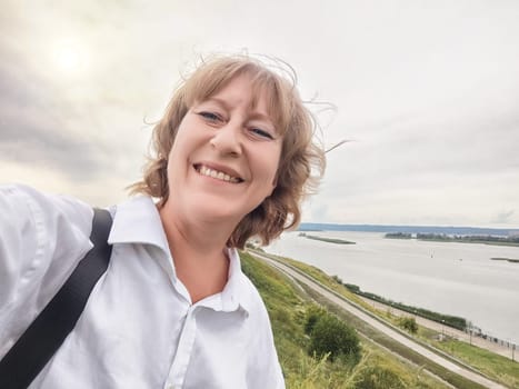 A middle-aged woman enjoys her time exploring nature while taking a selfie beside the river, smiling joyfully.
